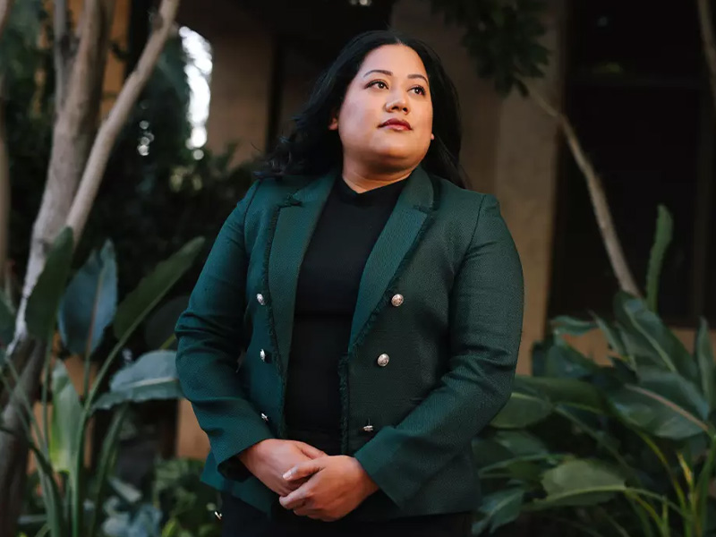 Jessie Lopez in a green coat looking off-camera in front of trees and green foliage.