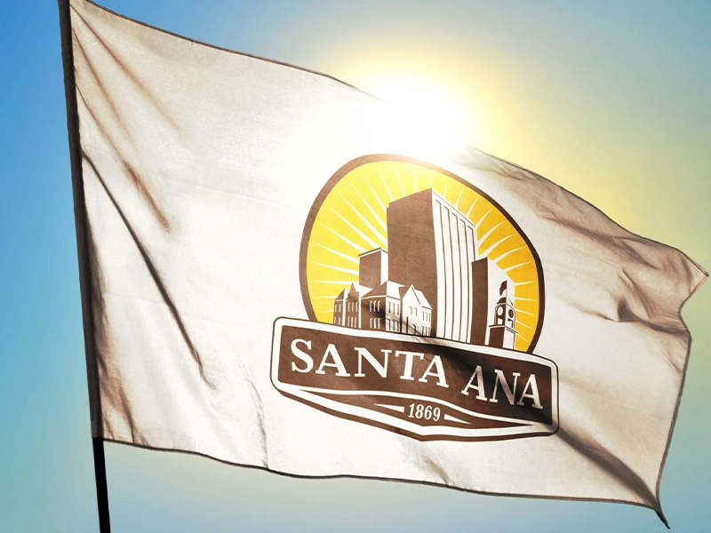 A flag with the Santa Ana logo on it waving in front of the sun and blue sky.