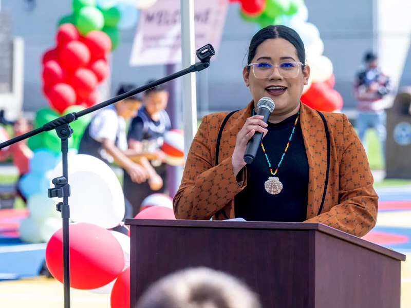 Jessie Lopez speaking at a podium in front of red, white, and green balloons.