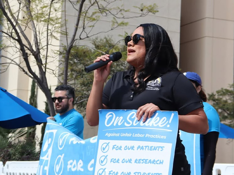 Councilmember Jessie Lopez speaking into a microphone and holding a blue "on strike" sign at the UPTE-CWA strike.