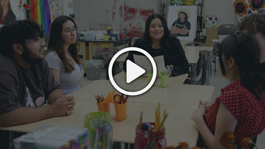 Thumbnail of Jessie Lopez sitting at a table in a colorful classroom with four teenagers, talking.