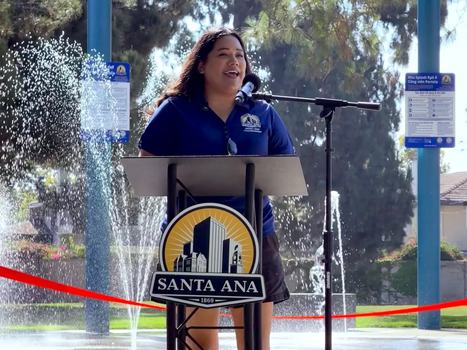 Jessie Lopez speaking at a podium in front of a new splash pad with water flaring.