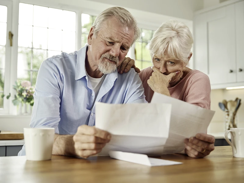 A married senior citizen couple looking distressed as they read two bills in their kitchen.