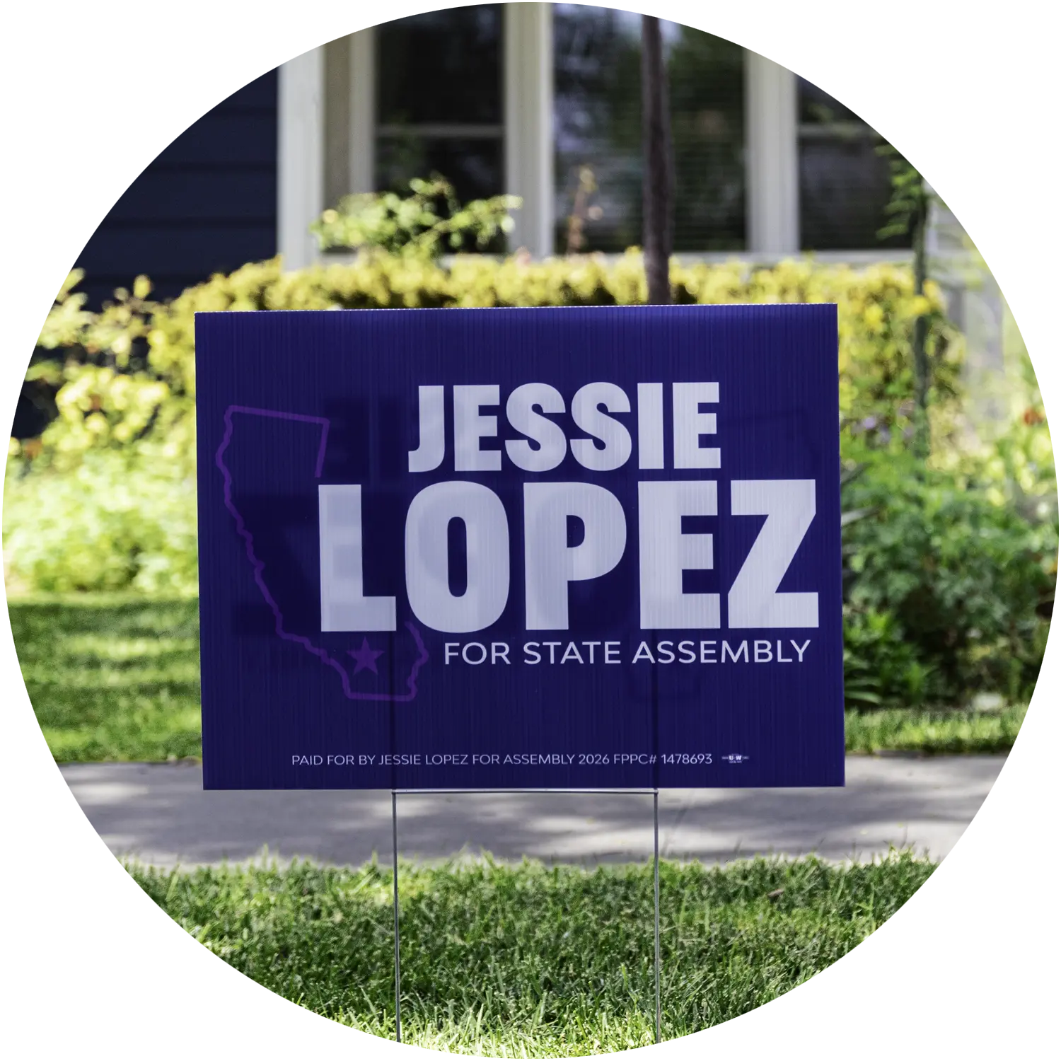A purple Jessie Lopez lawn sign displayed in front of a front yard and flowers.
