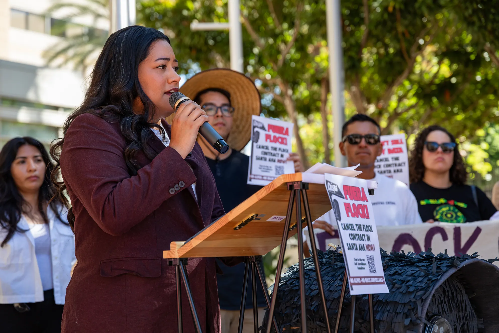 Jessie Lopez speaking into a microphone in front of individuals holding anti-Flock signs.